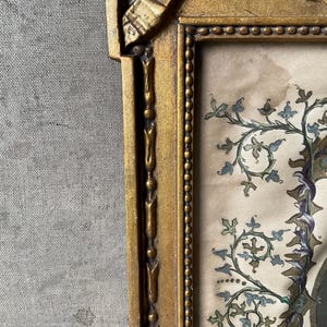 May include: A vintage framed artwork with a gold-colored ornate frame. The artwork features a detailed illustration with blue, green, and purple floral elements. The background is a light beige color.