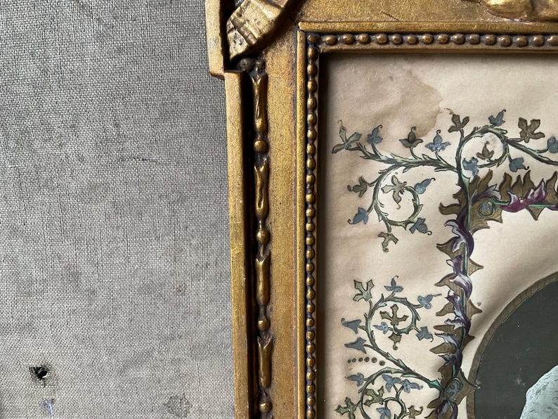 May include: A vintage framed artwork with a gold-colored ornate frame. The artwork features a detailed illustration with blue, green, and purple floral elements. The background is a light beige color.