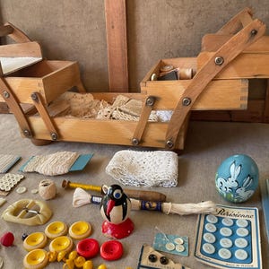 Op de afbeelding: Een open houten naaidoos met meerdere compartimenten, met spoelen, kant en andere naaispullen. Diverse vintage naaibenodigdheden zijn verspreid, waaronder knopen, een pinguïnbeeldje en een beschilderd ei met konijnen.