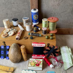May include: A collection of vintage sewing supplies including wooden spools of thread in various colors, a wooden box, a darning egg, a thimble, buttons, a measuring stick, and a red card labeled AU PEIGNE with black thread.