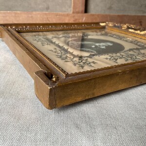 May include: A vintage, rectangular, gold-toned picture frame. The frame has a detailed, ornate design around the edges. The picture inside is a black and white image with a decorative border. The frame is resting on a light gray surface.