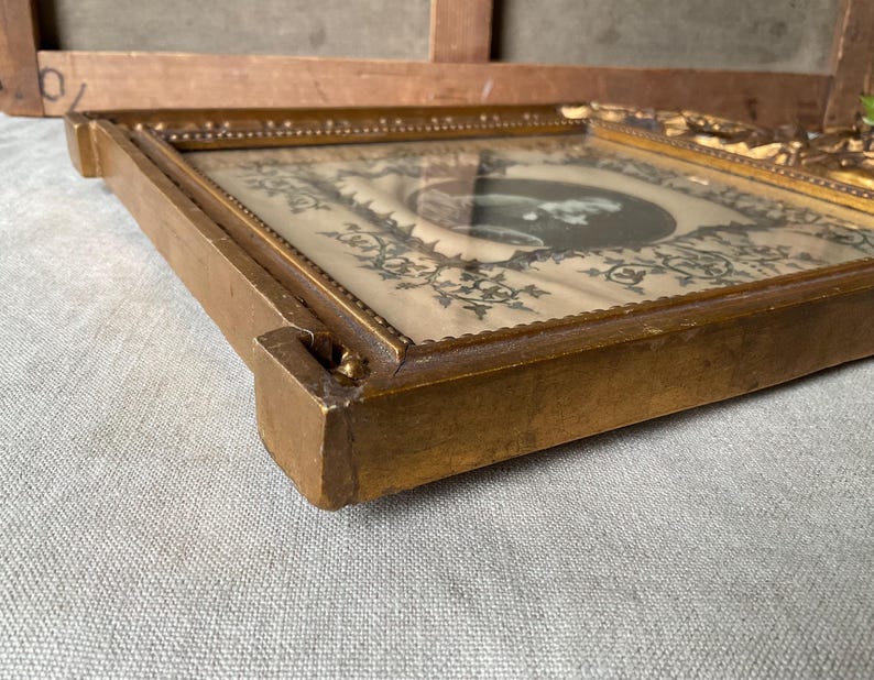 May include: A vintage, rectangular, gold-toned picture frame. The frame has a detailed, ornate design around the edges. The picture inside is a black and white image with a decorative border. The frame is resting on a light gray surface.