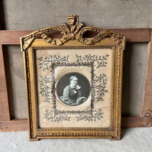 May include: Ornate gold-colored framed portrait. The frame has a decorative bow at the top and a rectangular shape. The portrait features a black and white image of a woman in a circle, surrounded by floral designs.