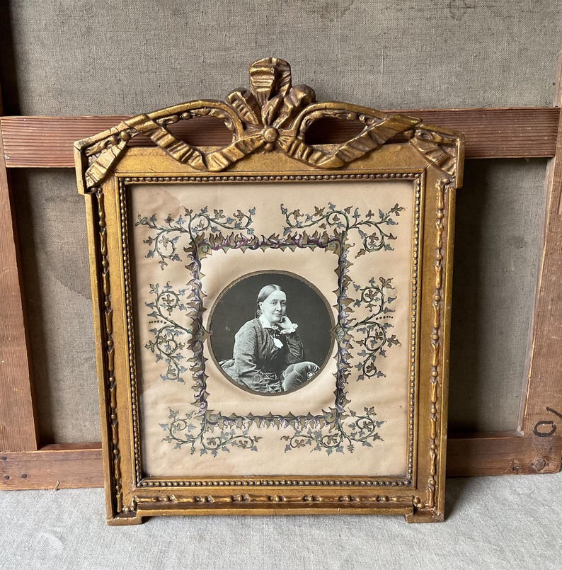 May include: Ornate gold-colored framed portrait. The frame has a decorative bow at the top and a rectangular shape. The portrait features a black and white image of a woman in a circle, surrounded by floral designs.