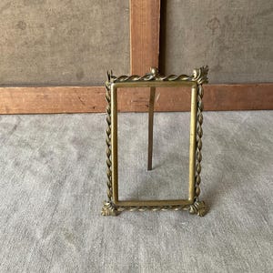 Puede incluir: Un marco de fotos de latón vintage con una abertura rectangular. El marco tiene un diseño de cuerda trenzada alrededor de los bordes y detalles florales decorativos en las esquinas. Un soporte metálico sostiene el marco.