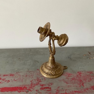 May include: Ornate gold-colored candlestick holder with a detailed, swirling design. The candle holder has a round base, a central stem, and two arms to hold candles. The design is vintage and decorative, suitable for home decor.