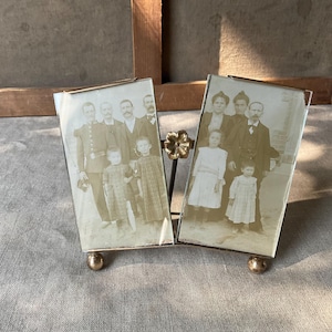 Puede incluir: Un marco de fotos doble vintage con dos retratos familiares en tonos sepia. El marco tiene un adorno floral dorado y descansa sobre dos pequeños pies esféricos dorados. Los retratos muestran grupos de personas con vestimenta formal.