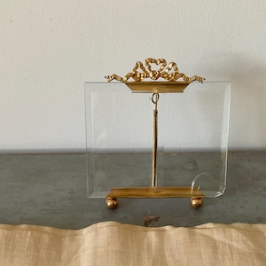 Può includere: Una cornice portafoto rettangolare in vetro con base in metallo dorato dotata di due piedini sferici e un'asta di supporto verticale centrale. La parte superiore della cornice è decorata con un elemento in metallo dorato a forma di fiocco.