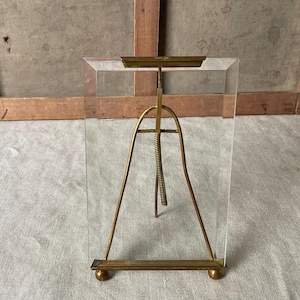 May include: A clear glass picture frame with a brass-coloured metal stand. The stand has a curved design with a central spring and adjustable top. The frame is rectangular and sits on two small ball feet.