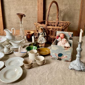 Op de afbeelding: Een verzameling vintage miniatuurvoorwerpen, waaronder een rieten mand, een kleine pop in een met stof beklede doos, een metalen kandelaar, diverse keramische borden, kopjes, een sauskom, een kan, een glazen fles met trechter en een bruine kruik.