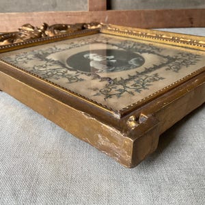May include: A vintage, square-shaped picture frame with a gold finish. The frame holds a black and white portrait surrounded by floral and vine designs. The frame's edges are ornate, and the overall aesthetic is antique.