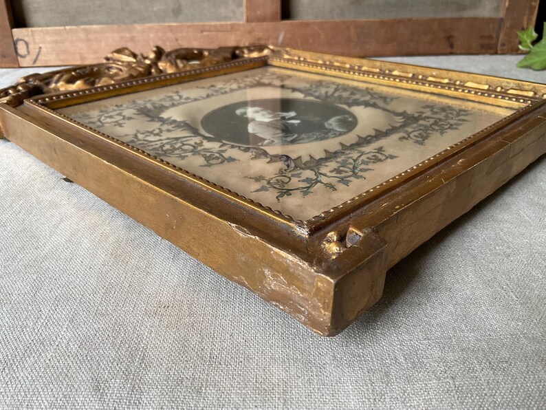 May include: A vintage, square-shaped picture frame with a gold finish. The frame holds a black and white portrait surrounded by floral and vine designs. The frame's edges are ornate, and the overall aesthetic is antique.