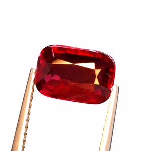 Könnte beinhalten: Ein facettierter, rechteckig geschliffener Edelstein, der von einer Pinzette gehalten wird. Der Edelstein hat eine tiefrote Farbe mit inneren Lichtreflexen. Die Pinzette ist goldfarben, und der Hintergrund ist weiß.