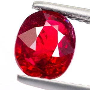 Könnte beinhalten: Ein facettierter, oval geformter Rubin-Edelstein, der von einer silbernen Pinzette gehalten wird. Der Rubin hat eine tiefe, leuchtende rote Farbe mit mehreren Facetten, die das Licht reflektieren. Der Edelstein ist vor weißem Hintergrund.