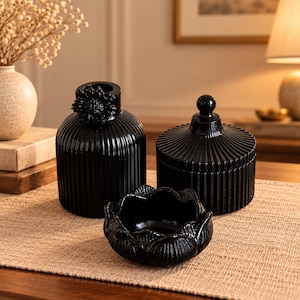 Può includere: Un set di tre oggetti decorativi in vetro nero composto da un vaso a coste con un dettaglio floreale, un barattolo con coperchio dalla texture a coste e pomello rotondo, e una piccola ciotola a forma di fiore con petali curvi.