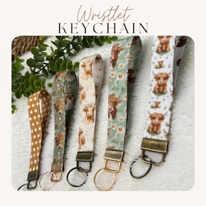 May include: Five wristlet keychains with various patterns, including polka dots, Highland cows, and floral designs. The keychains have metal hardware and key rings. The text "Wristlet KEYCHAIN" is at the top of the image.