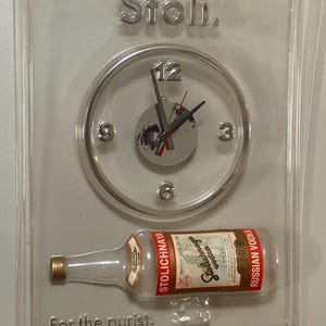 以下が含まれることがあります： 透明なアクリル製の壁掛け時計。Stoliウォッカのボトルが特徴です。時計は透明な文字盤で、黒い針があり、文字盤の上には「Stoli.」の文字があります。ボトルには「Stolichnaya Russian Vodka」と書かれており、その下には「For the purist.」の文字があります。
