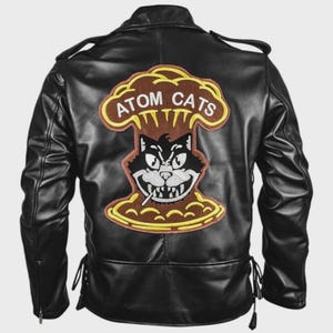 Può includere: Giacca in pelle nera con una grande toppa sul retro con la scritta "ATOM CATS" in lettere gialle. La toppa raffigura un gatto dei cartoni animati che fuma una sigaretta, su uno sfondo a forma di fungo atomico. La giacca ha un classico stile biker.