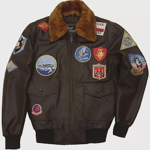 May include: A brown leather bomber jacket with a faux fur collar and multiple embroidered patches. The jacket features a zip-up front, two front pockets, and black ribbed cuffs and hem. Patches include military and state emblems.
