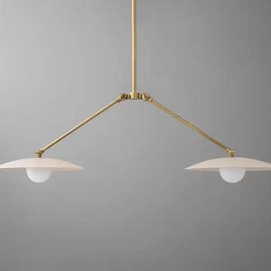 Könnte beinhalten: Moderne Pendelleuchte mit zwei Lichtern und Messing-Finish. Sie hat zwei cremefarbene, scheibenförmige Schirme, jeweils mit einer weißen Glühbirne. Die Arme erstrecken sich von einer zentralen Stange.