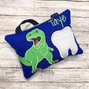 May include: Blue tooth pillow with a green dinosaur and a white tooth embroidered on it. The pillow has a black handle and the name "Taye" is embroidered on the top.