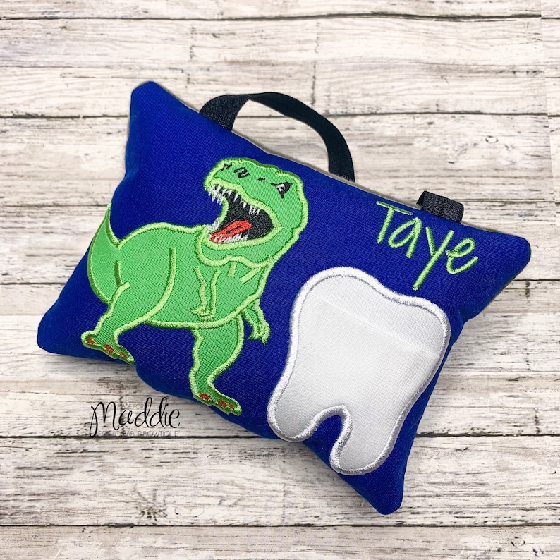 May include: Blue tooth pillow with a green dinosaur and a white tooth. The pillow has a black handle and the name "Taye" is embroidered on it.