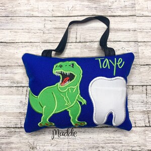May include: Blue tooth pillow with a green dinosaur and a white tooth. The pillow has a black loop for hanging and the name "Taye" is embroidered on the pillow.