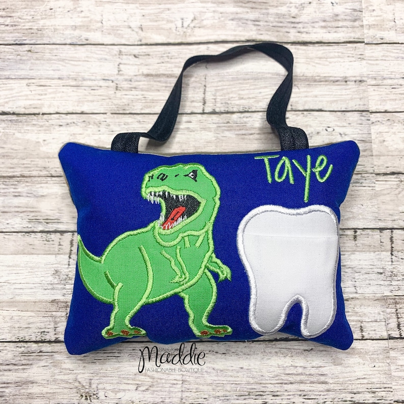 May include: Blue tooth pillow with a green dinosaur and a white tooth. The pillow has a black loop for hanging and the name "Taye" is embroidered on the pillow.