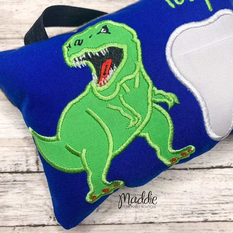 May include: A blue pillow with a green dinosaur and a white tooth embroidered on it. The pillow has a black handle and the text "Made for Maddie Fashionable Boutique" is embroidered on the bottom.