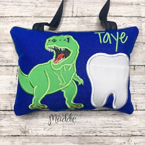 May include: A blue pillow with a green dinosaur and a white tooth embroidered on it. The pillow has a black strap and the name "Taye" is embroidered in green on the top right corner.