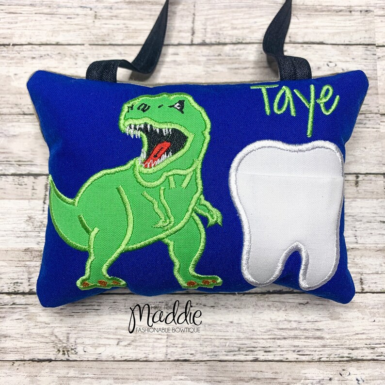 May include: A blue pillow with a green dinosaur and a white tooth embroidered on it. The pillow has a black strap and the name "Taye" is embroidered in green on the top right corner.