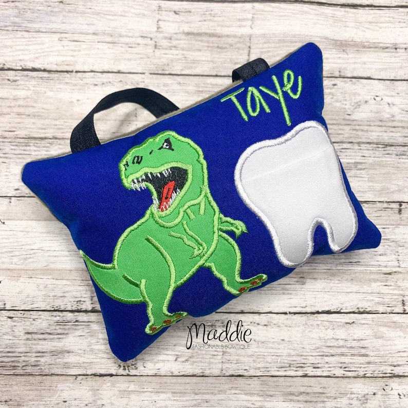 May include: Blue tooth pillow with a green dinosaur and a white tooth embroidered on it. The pillow has a black handle and the name "Taye" is embroidered on the top.