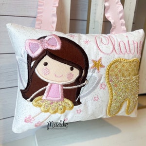 May include: A white pillow with a pink tooth fairy embroidered on it. The fairy has brown hair, a pink dress, and a gold star wand. The pillow also has a gold tooth and the name "Claire" embroidered on it.