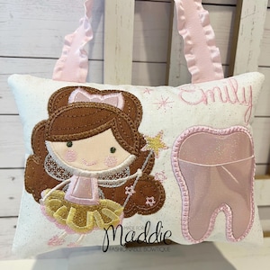 May include: A white pillow with a pink tooth pocket and a pink fairy with brown hair and a gold skirt. The pillow has the name "Emily" embroidered on it and the text "Made for Maddie Fashionable Bowtique".