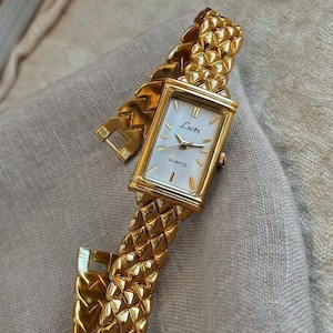 May include: A gold-toned wristwatch with a rectangular face and a textured band. The watch face is white with black hands and the word "Lutr" printed on it. The band is made of interlocking geometric shapes.