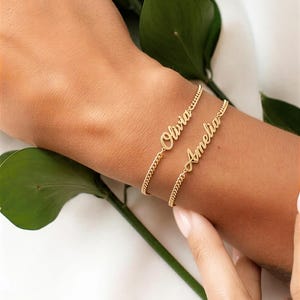 May include: Two gold-colored chain bracelets, each with a name in a cursive font. The top bracelet says "Olivia" and the bottom bracelet says "Amelia". The bracelets are on a person's wrist, with a green leaf in the background.