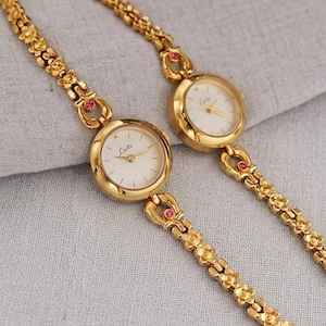 Puede incluir: Dos relojes de pulsera dorados con esferas blancas y delicadas correas florales. Las esferas muestran las marcas "Lucte" y "Cove". Cada reloj tiene una esfera pequeña y redonda y está adornado con una pequeña gema roja.