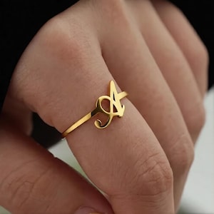 Può includere: Un anello dorato con la lettera "A" in un delicato carattere corsivo. L'anello è sottile e minimalista, con una finitura lucida. Viene indossato su un dito, mostrando il suo design elegante.
