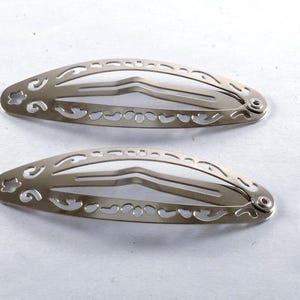 May include: Two silver-colored hair barrettes. Each barrette has an oval shape with decorative cut-out patterns along the edges. The barrettes are made of a shiny metal and are lying flat on a white surface.