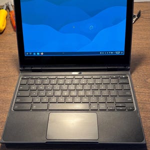 May include: A black Lenovo laptop with a blue desktop background featuring a wave pattern. The device has a standard QWERTY keyboard, a trackpad, and a screen displaying a taskbar with various icons, including a web browser and system settings.