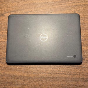 May include: A closed, dark gray Dell Chromebook laptop on a wooden surface. The Dell logo is visible in the center of the lid. The word "chrome" is printed on the lower right corner of the laptop.