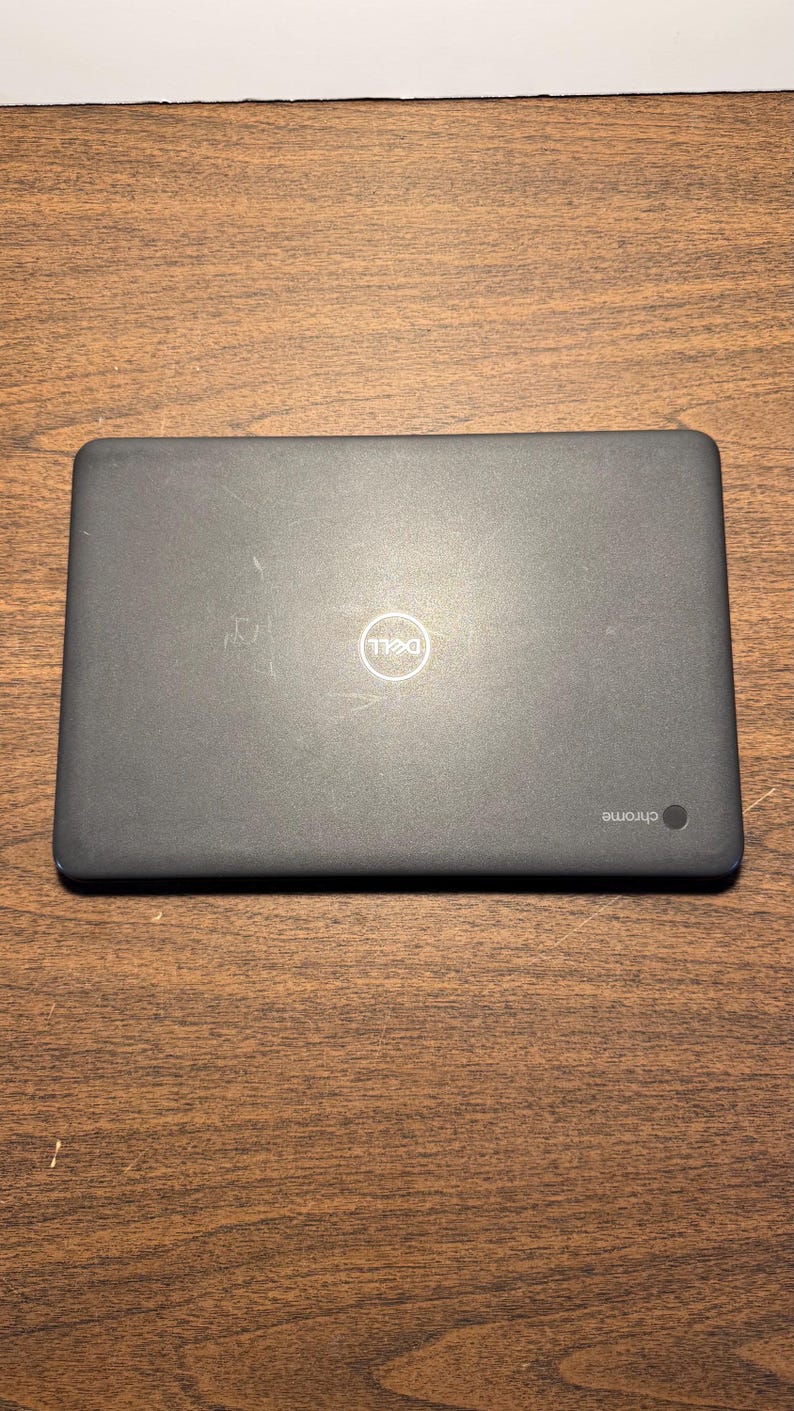 May include: A closed, dark gray Dell Chromebook laptop on a wooden surface. The Dell logo is visible in the center of the lid. The word "chrome" is printed on the lower right corner of the laptop.