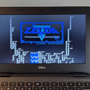 May include: A black Dell laptop displaying the title screen of the 1986 Nintendo game "The Legend of Zelda." The screen shows the game's logo in blue and white pixelated graphics. The laptop keyboard is visible below the screen.