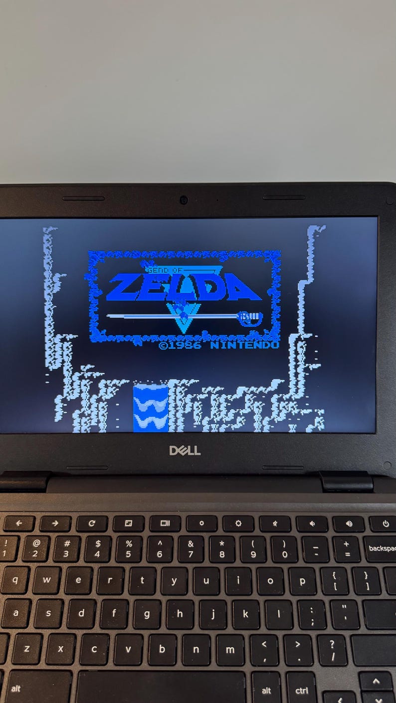 May include: A black Dell laptop displaying the title screen of the 1986 Nintendo game "The Legend of Zelda." The screen shows the game's logo in blue and white pixelated graphics. The laptop keyboard is visible below the screen.
