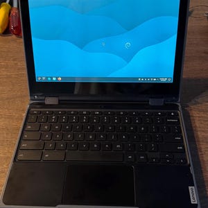 May include: A Lenovo laptop with a dark gray chassis, a black keyboard, and a trackpad. The screen displays a blue desktop background with a light blue wave pattern and a taskbar at the bottom containing various icons and a system clock.