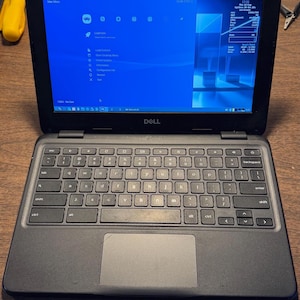 May include: A black Dell laptop with a QWERTY keyboard and a trackpad. The screen displays a blue interface with the RetroArch menu, system information, and a taskbar at the bottom. The device features a webcam at the top center of the bezel.