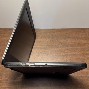 May include: A black laptop computer with the screen open. The laptop is on a wooden surface. The side profile shows a USB port, a charging port, and a headphone jack.