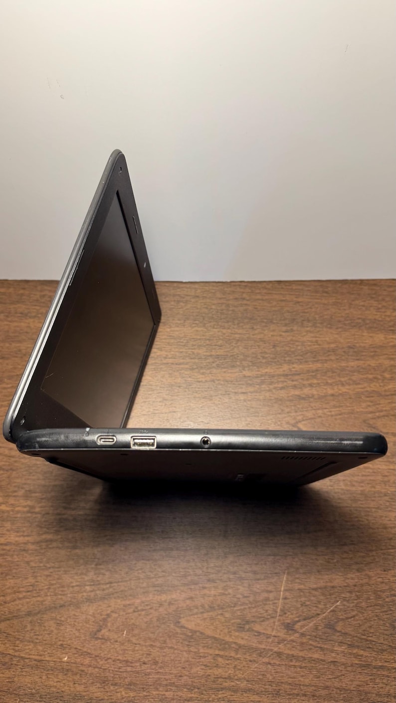 May include: A black laptop computer with the screen open. The laptop is on a wooden surface. The side profile shows a USB port, a charging port, and a headphone jack.