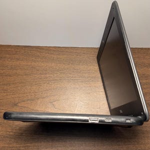 May include: A black laptop computer with a silver trim is open on a wooden surface. The screen is dark, and the side profile shows a USB port and other connectors. The laptop is angled, displaying its screen.