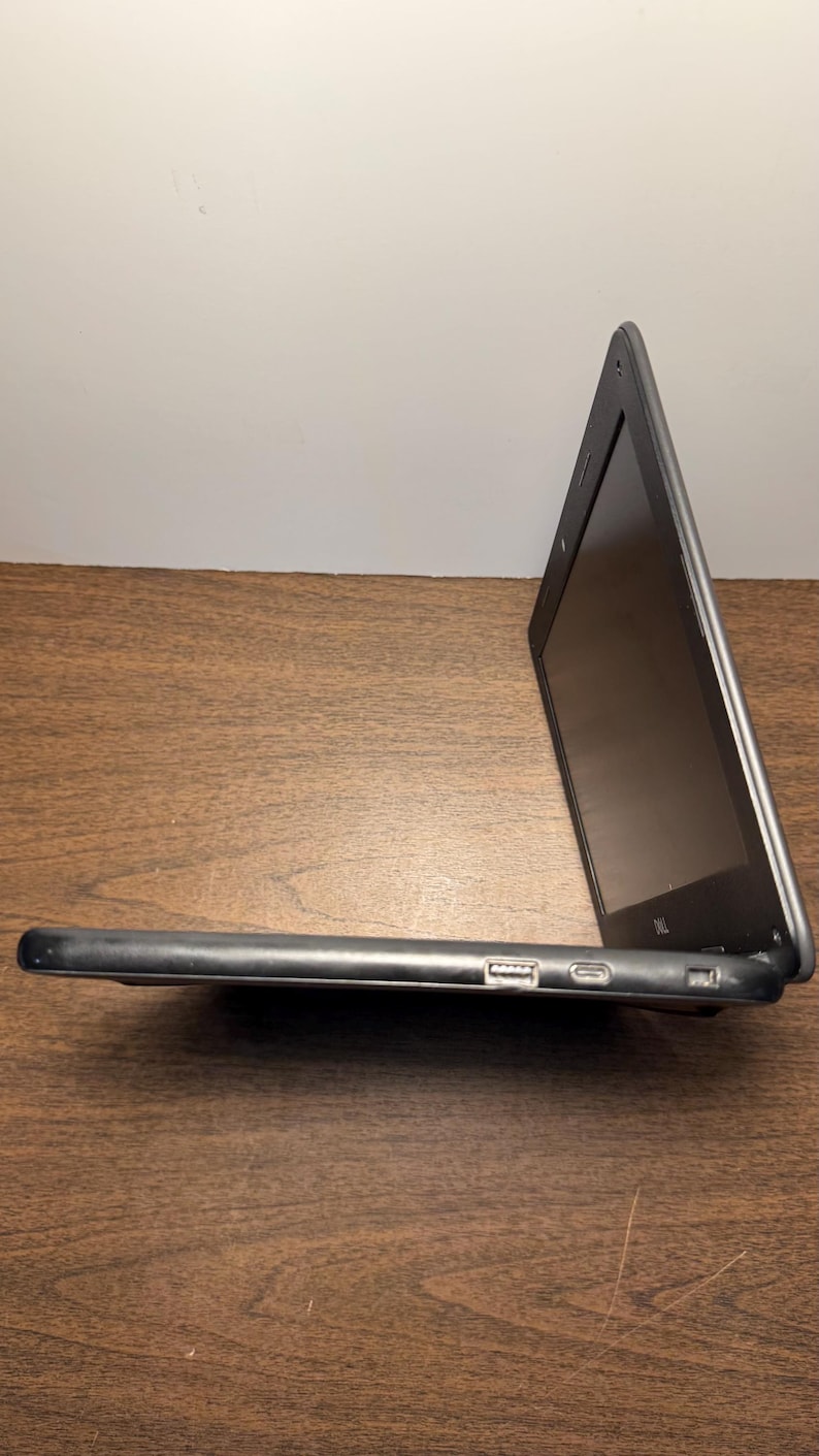 May include: A black laptop computer with a silver trim is open on a wooden surface. The screen is dark, and the side profile shows a USB port and other connectors. The laptop is angled, displaying its screen.